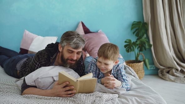 Caring Father Is Reading Funny Book To His Child While Boy Is Laughing and Talking To His Parent