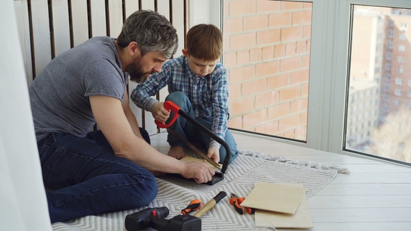 Little Boy Is Focused on Sawing Piece of Wood with Hand Saw with His Father Helping and Teaching Him