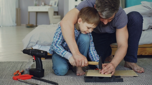 Father Bearded Man Is Teaching His Son How To Use Hammer Driving Nail in Piece of Wood Together