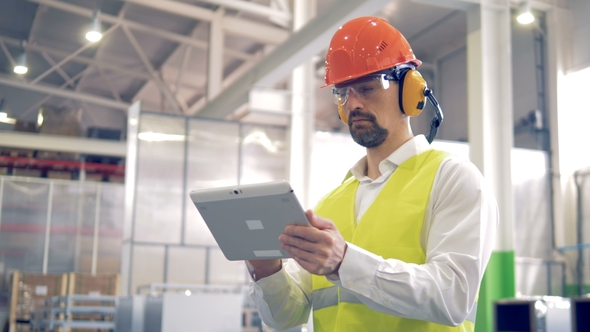 Factory Worker Is Standing and Working with His Tablet at a Factory Warehouse