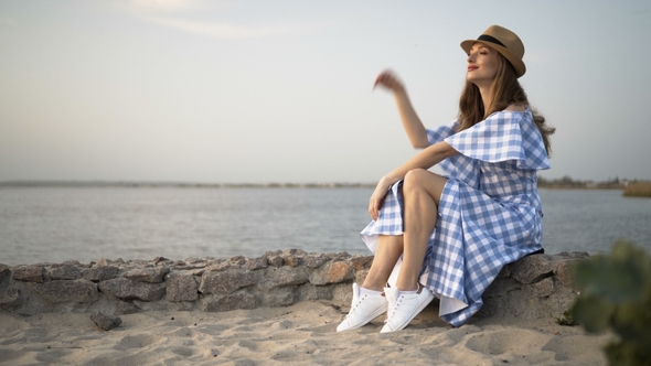Attractive Woman in Hat and Dress Is Sitting and Looking at Sunset