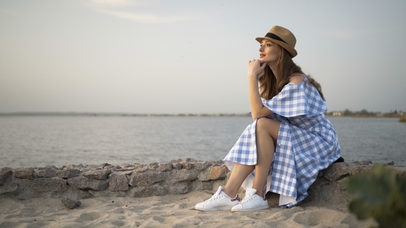 Attractive Woman in Hat and Dress Is Sitting and Looking at Sunset