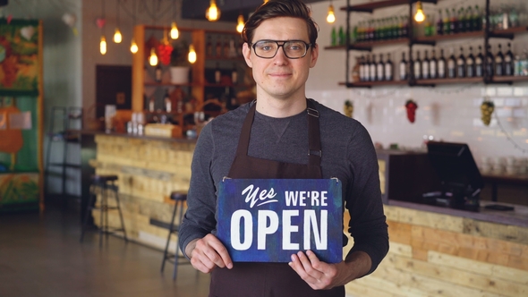 Cheerful Handsome Young Waiter in Apron Holding "We Are Open" Sign Standing in Opening