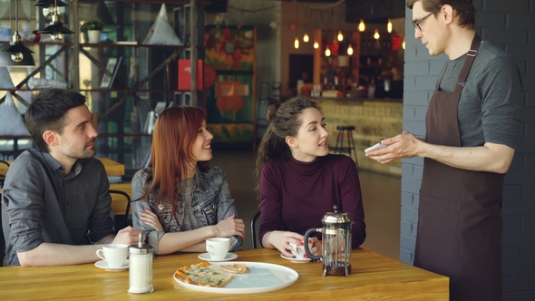 Friendly Male Waiter Taking Order from Cheerful Group of Friends Sitting at Table in Cafe