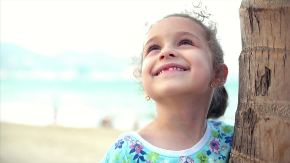 Happy Little Girl on the Beach in a Blue Dress with a Palm Tree, Looking at the Camera, Smiling and