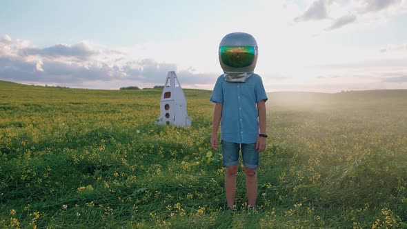 Dreamer Boy in an Astronaut Helmet Standing in a Field of Rapeseed Near Cardboard Model of a Space