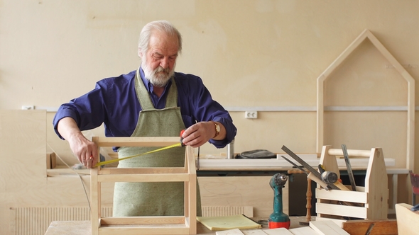 Old Grey Haired Carpenter with Moustache and Beard Measuring Wooden Doll House.