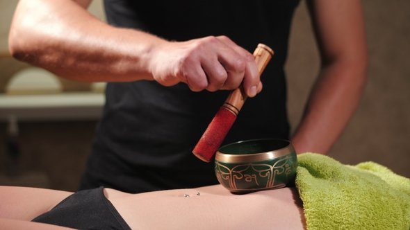 Male Masseuse Plays on the Singing Bowl on Woman's Belly in Spa Center