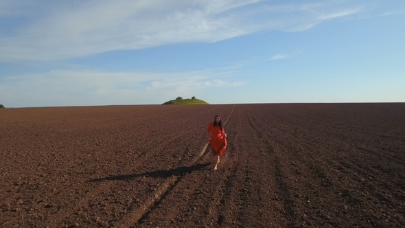 Charming Brunette Wearing Long Red Dress Walking at Plowed Field