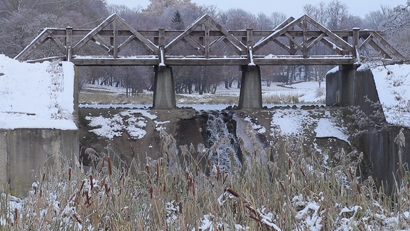 Little Waterfall Under the Bridge on the Background of Lake and Some Snowfall