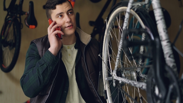 Serious Young Man Bike Repairman Is Checking and Turning Wheel While Talking on Mobile Phone