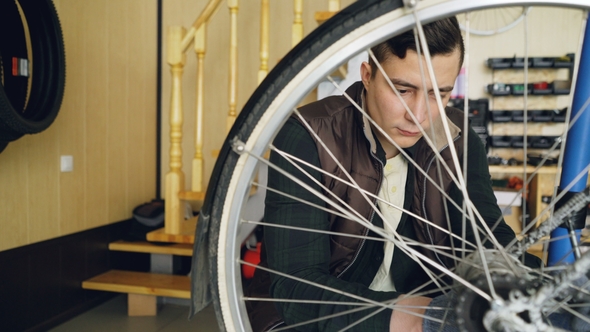 Young Man Experienced Serviceman Is Fixing Bike Wheel Using Wrench and Tools. Small Cozy Workshop