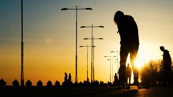 Silhouettes of Young Guys Riding on Bicycles, Skateboards, Roller Skates in a Park at Sunset