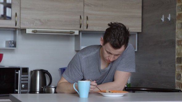 Sleepy Young Man Eating Oat Breakfast Cereal in the Kitchen