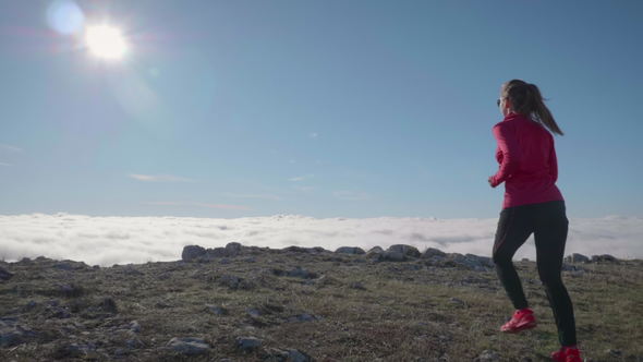 Young Caucasian Woman in Sunglasses Is Jogging in Mountains Above Clouds at Sunny Day. Sky Running