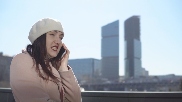 Woman in Coat with Phone Against a Background of High Buildings of a Megacity
