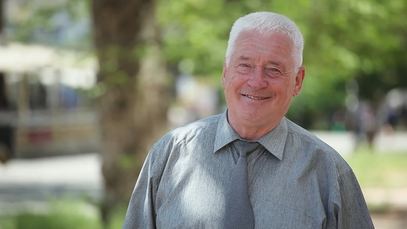 Kind White-haired Man Stands in a Green Street and Smiles on a Sunny Day