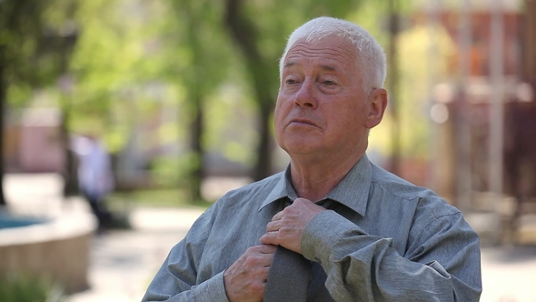 Confident Old Man Pulls Up His Tie While Waiting a Lady in a Street in Summer