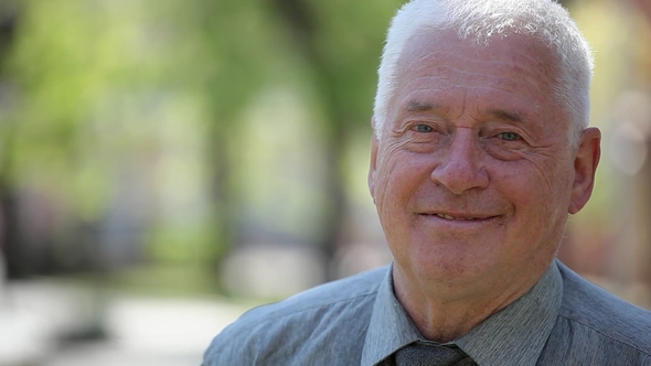 Happy White-haired Man Stands in a Green Street and Smiles on a Sunny Day