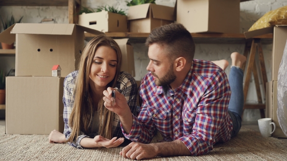 Young Cute Couple in Casual Clothes Is Lying on Floor of New Apartment, Guy Is Giving Keys