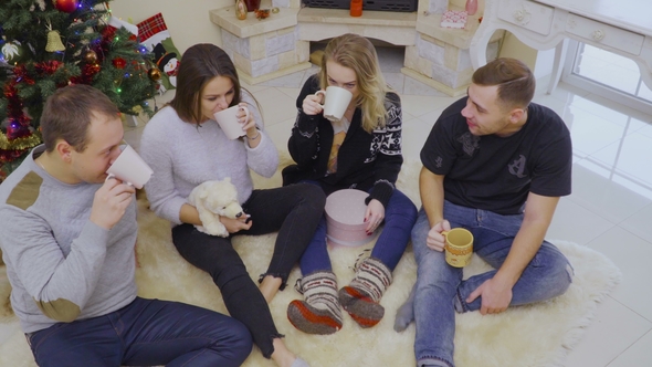 Four Young People Drinks Tea or Coffee Sitting on Floor Near Christmas Tree
