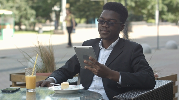 Handsome Afro American Man Is Using a Tablet, While Sitting in Outside Cafe