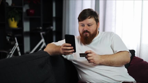 Serious Fat Man Types a Number of His Credit Card in the Smartphone Sitting on the Couch