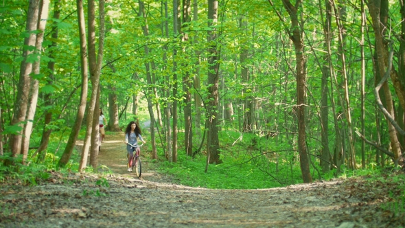 Two Girls on Bicycles Climb the Path to the Hill in Green Forest