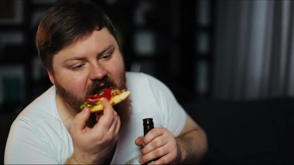 Fat Man with Beard Eats Pizza Sitting at the Table with Beer and Pop-corn Before a TV-set