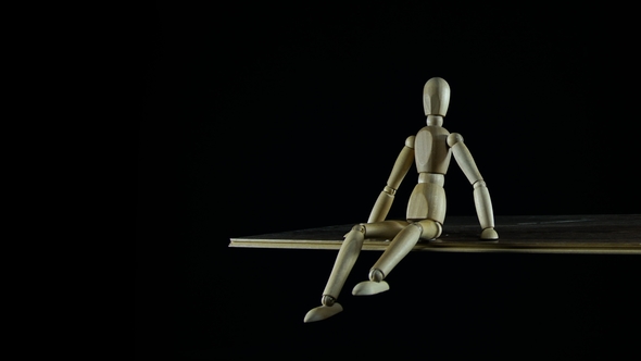 Wooden Figure Dummy Procrastinates in Studio on Black Background Sitting and Waving Legs