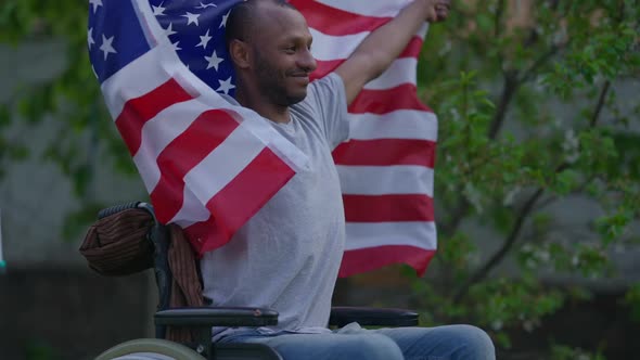 Satisfied Smiling African American Man in Wheelchair Fluttering USA Flag in Hands Looking Away