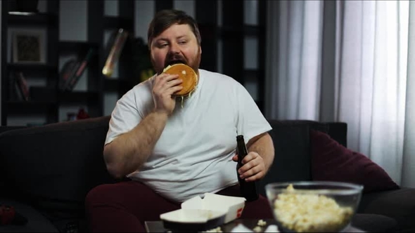 Fat Man Looks at the TV-set Drinking Beer and Eating a Burger