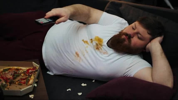 Fat Man in Dirty T-shirt Laughs Watching the TV-set on His Couch in a Living Room