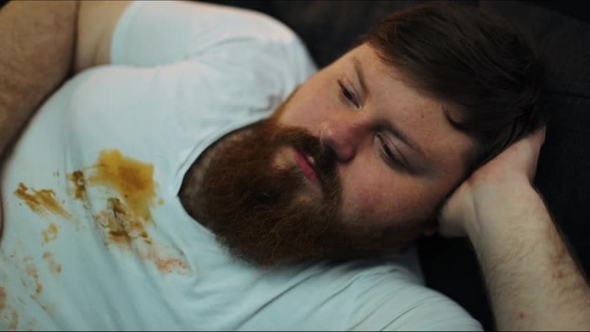 Fat Man in Dirty T-shirt Laughs Watching the TV-set on His Couch in a Living Room