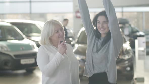 Happy Young Brunette Woman Taking Car Keys From Positive Mature Mother, Dancing and Hugging Parent