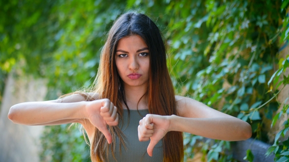 Young Beautiful Woman Standing on Green Foliage Background Expressing Discontent and Showing Thumb