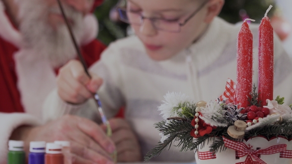 Santa and Little Boy Draw Picture at Blurred Background of Two Festive Candles
