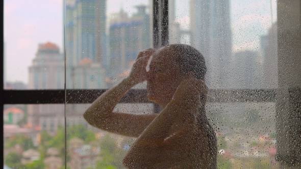 Young Woman Takes a Shower in the Bathroom with Panoramic Windows and Silhouettes of Skyscrapers