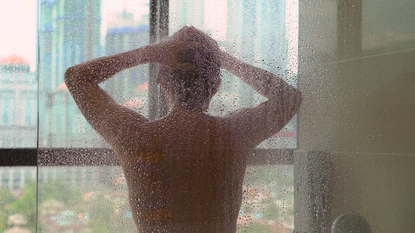 Young Man Takes a Shower in the Bathroom with Panoramic Windows and Silhouettes of Skyscrapers