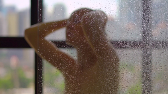 Young Woman Takes a Shower in the Bathroom with Panoramic Windows and Silhouettes of Skyscrapers