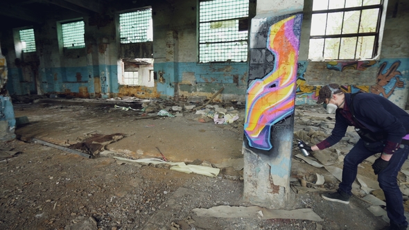 Pan Shot of Masked Graffiti Artist Drawing Abstract Images on Pillar in Large Empty Building Using