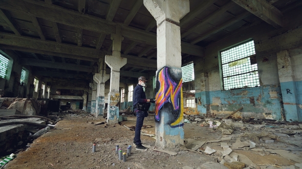 Graffiti Artist in Protective Mask Is Painting on High Column in Abandoned Industrial Building