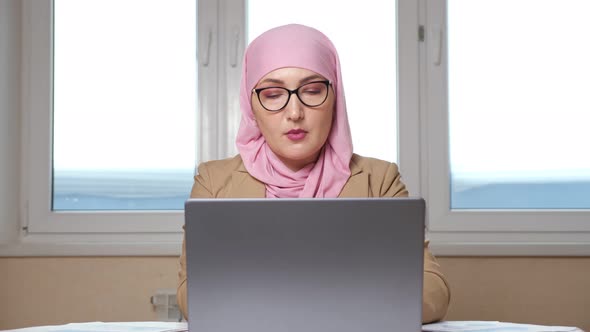 Muslim Woman in Pink Headscarf and Glasses Working on Laptop and with Documents