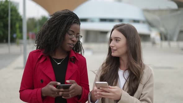 Two Women Walking Along Street, One Showing Pictures on Phone