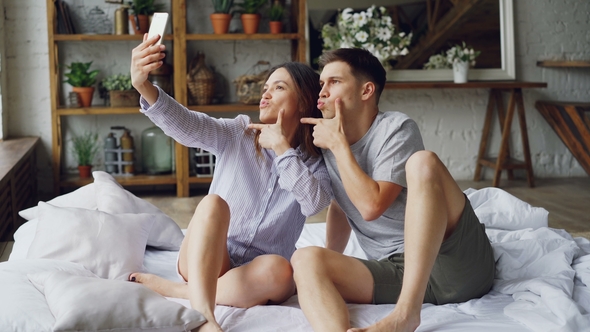 Modern Married Couple Is Taking Selfie in Bedroom Gesturing Posing and Kissing While Sitting on Bed