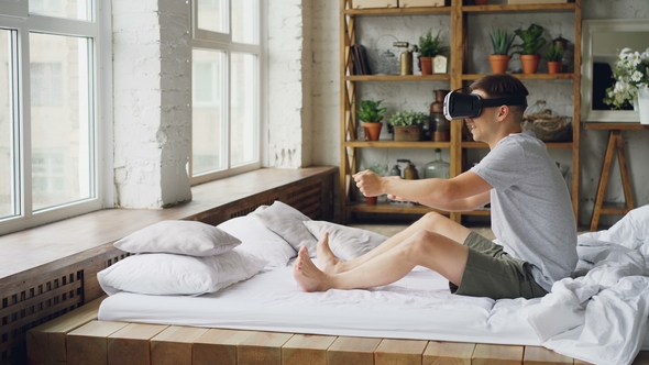 Happy Young Man in Virtual Reality Glasses Is Sitting on Bed and Moving Hands As If He Is Driving