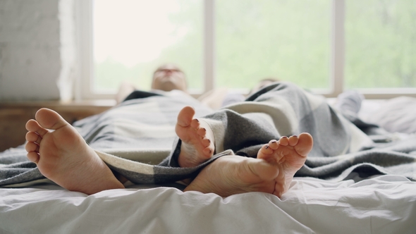 Adorable Loving Couple Is Lying in Bed Under Blanket and Talking, Focus on Bare Feet Touching