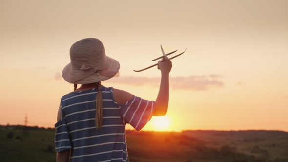 A Girl with Pigtails and a Hat Playing with a Wooden Airplane at Sunset. The Dream of Long-distance