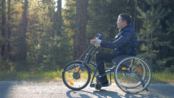 A Handicapped Man Goes on a Wheelchair on a Forest Road, Side View