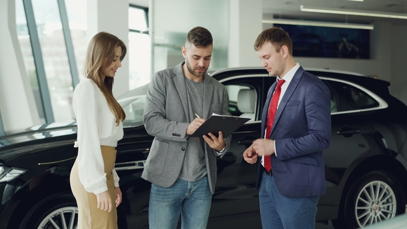 Handsome Young Guy Is Signing Sale and Purchase Agreement in Car Dealership